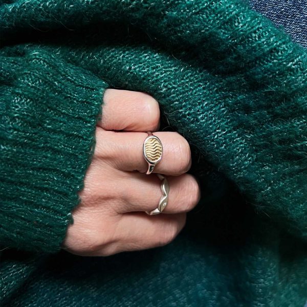 Jeune femme portant un pull vert montrant sa main avec des bagues artisanales : une bague type chevalière avec un détail ancien serti et des bagues en argent avec brillant.