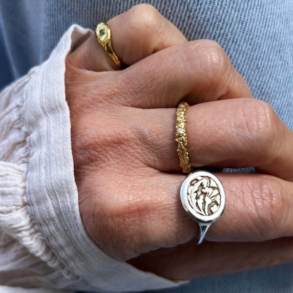 Femme portant une chemise blanche et un jean montrant sa main avec des bagues artisanales : une bague type chevalière avec motif figuratif serti, une bague organique avec brillant et une autre bague en or au petit doigt avec une émeraude verte.