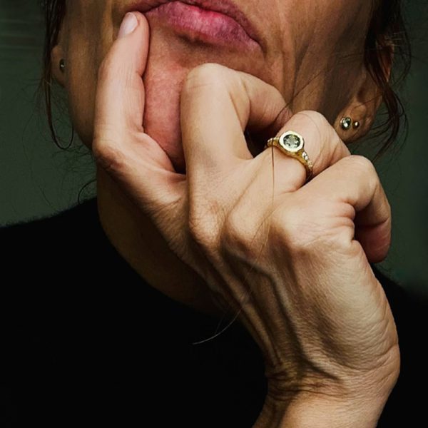 Mano femenina con anillo de oro reciclado de 18k hecho a medida, diseño artesanal con zafiro verde central y ocho diamantes blancos, acabado satinado-brillante y forma orgánica.
