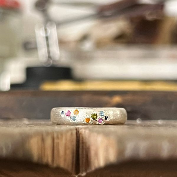 Bague en or blanc recyclé faite sur mesure, forme large et organique, sertie de saphirs verts, roses, jaunes, bleu Ceylan et Royal, finition satinée rustique.