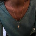 Collier long en or avec pendentif rond gravé de fleurs anciennes, porté sur un t-shirt vert à col en V
