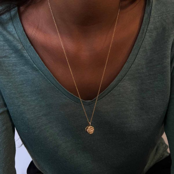 Collier long en or avec pendentif rond gravé de fleurs anciennes, porté sur un t-shirt vert à col en V