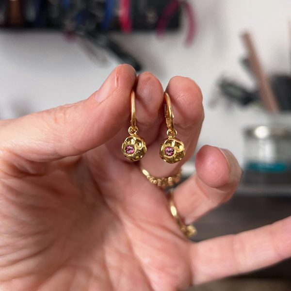 Boucles d’oreilles en or 18k martelé serties d’une tourmaline rose, tenues à la main sur fond flou.